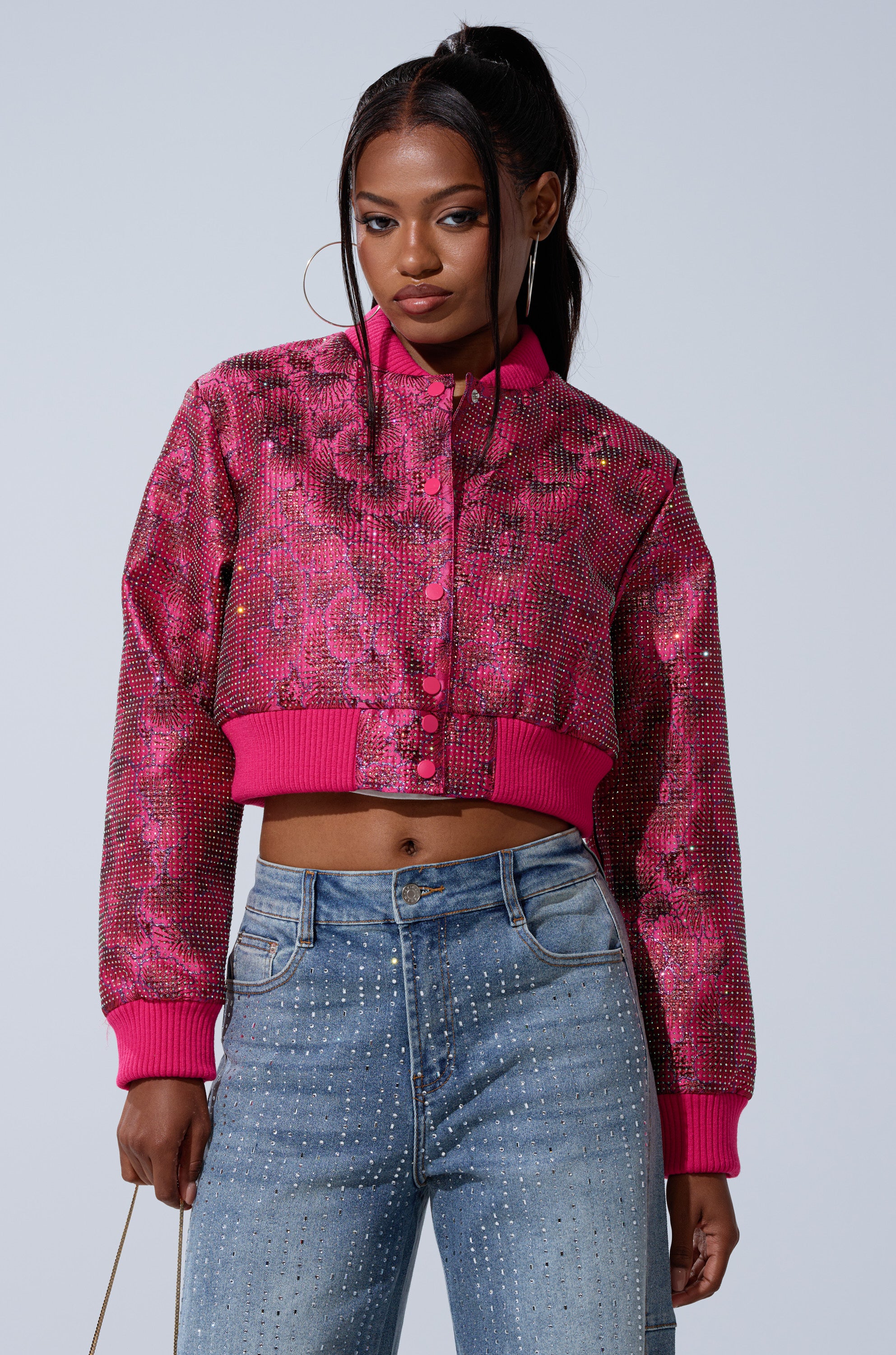 A woman poses confidently against a plain background, wearing the FLEUR RHINESTONE BROCADE BOMBER—a bright pink sequin-embellished cropped jacket with ribbed cuffs and collar—paired with high-waisted blue rhinestone jeans.