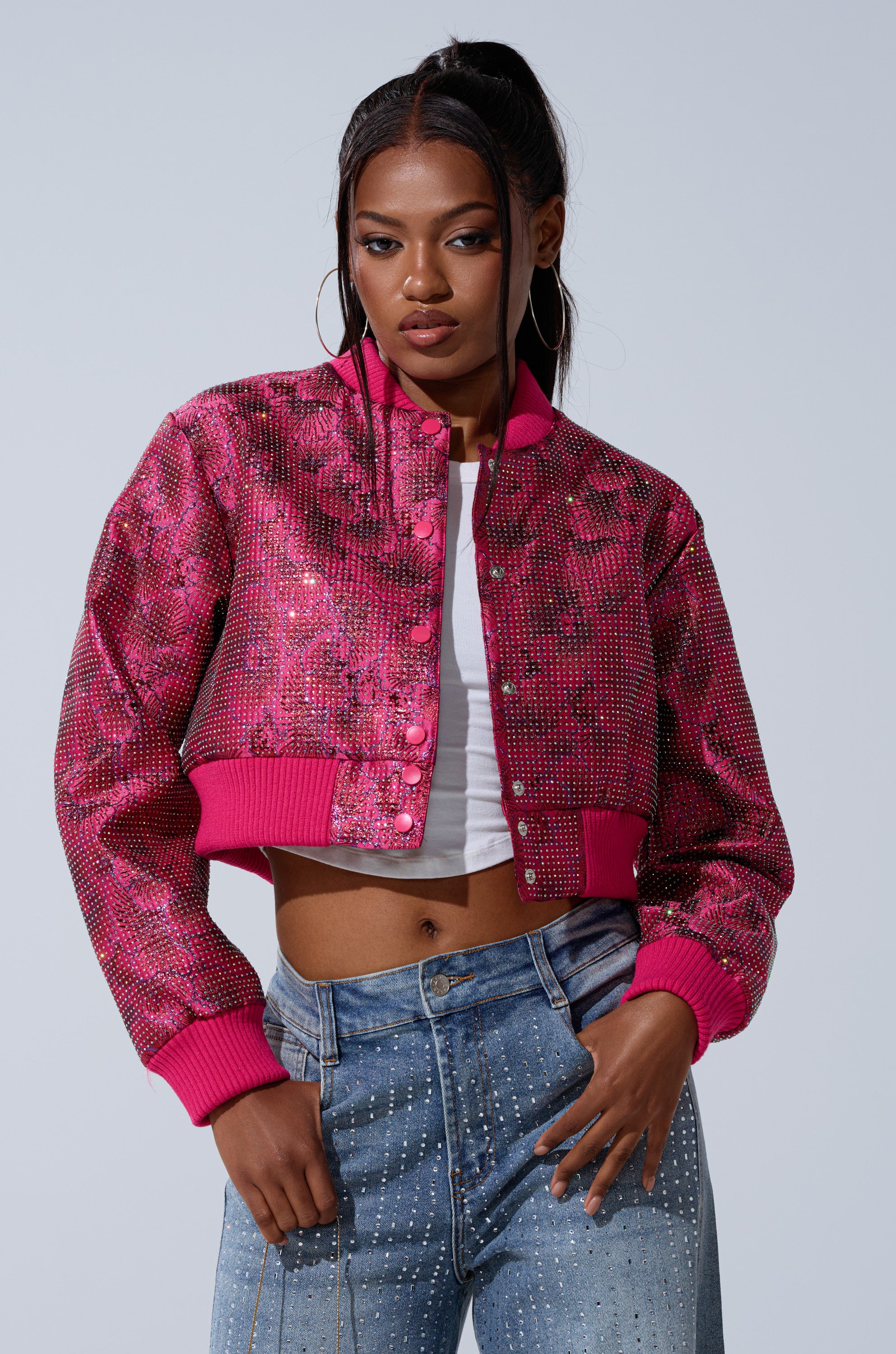 A woman with long dark hair in a high ponytail poses confidently against a plain background, wearing the FLEUR RHINESTONE BROCADE BOMBER over a white top and blue jeans with rhinestone embellishments.