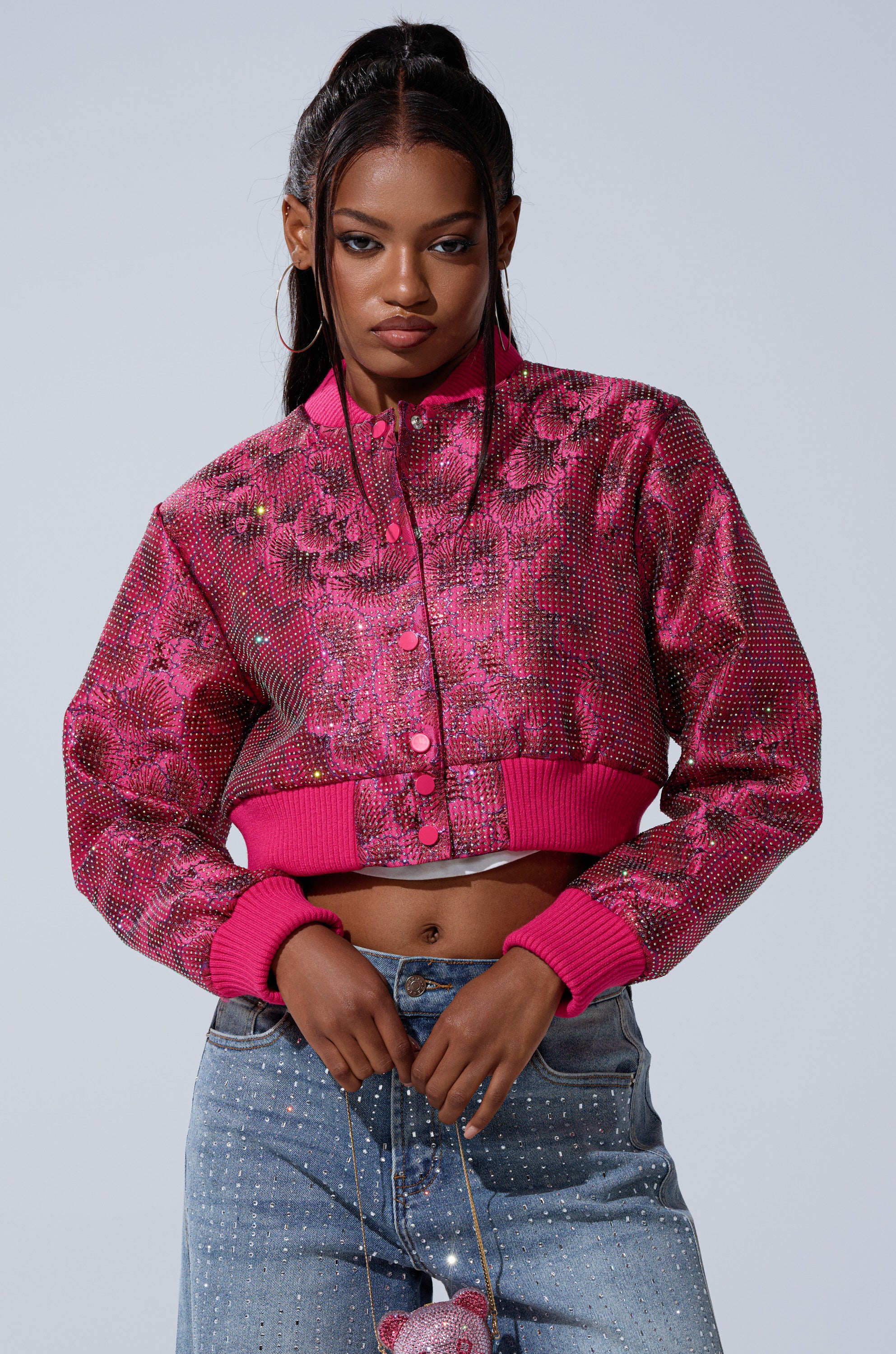 A woman wears the FLEUR RHINESTONE BROCADE BOMBER—bright pink and floral—with light blue rhinestone jeans, confidently standing with hands on her waist and looking at the camera.