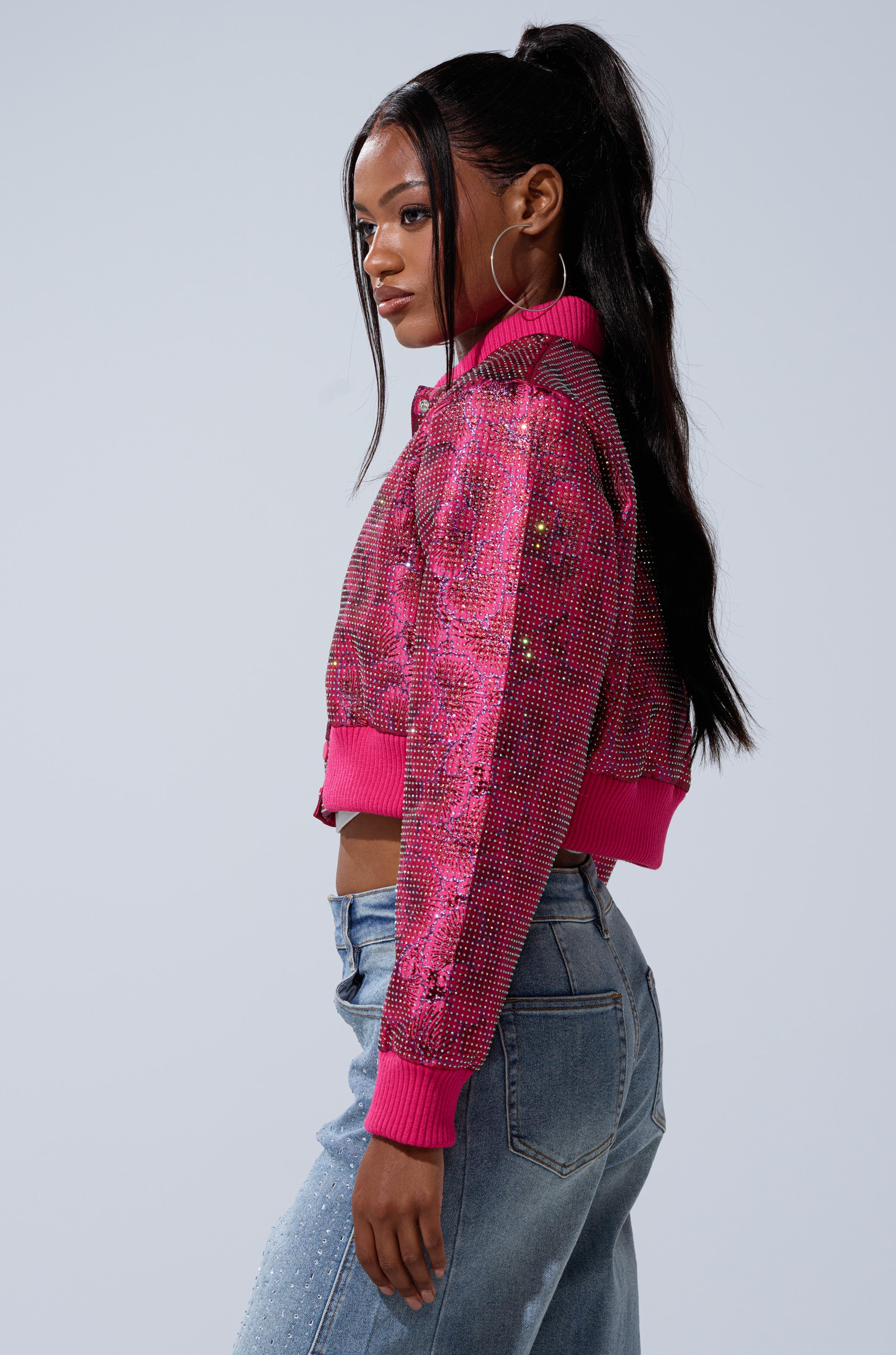 A woman with long black hair in a high ponytail models the FLEUR RHINESTONE BROCADE BOMBER, paired with light blue jeans, posing sideways against a plain light background.