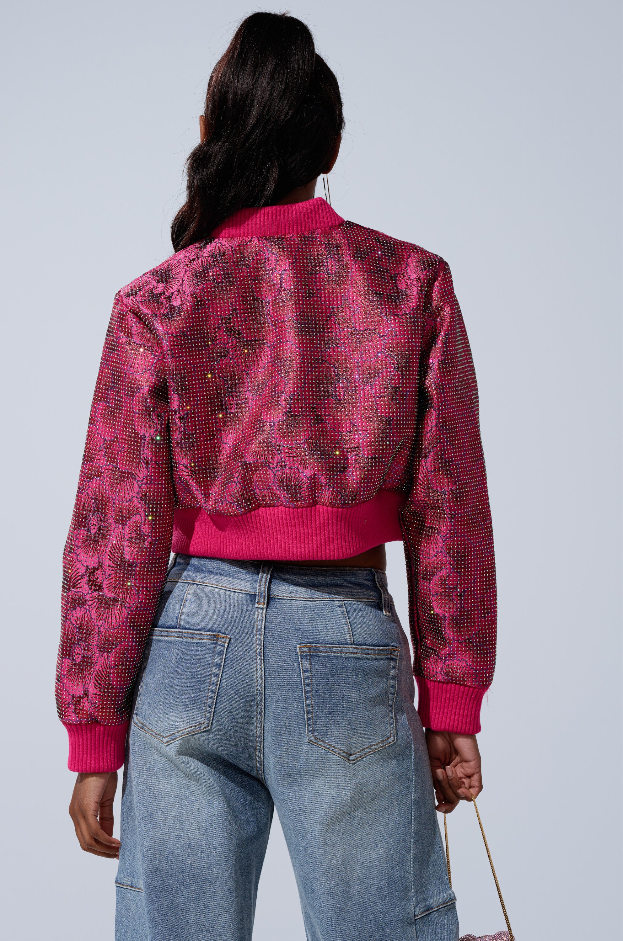 A person with long dark hair in a ponytail is seen from the back wearing the FLEUR RHINESTONE BROCADE BOMBER and light blue high-waisted jeans, set against a plain light gray background.