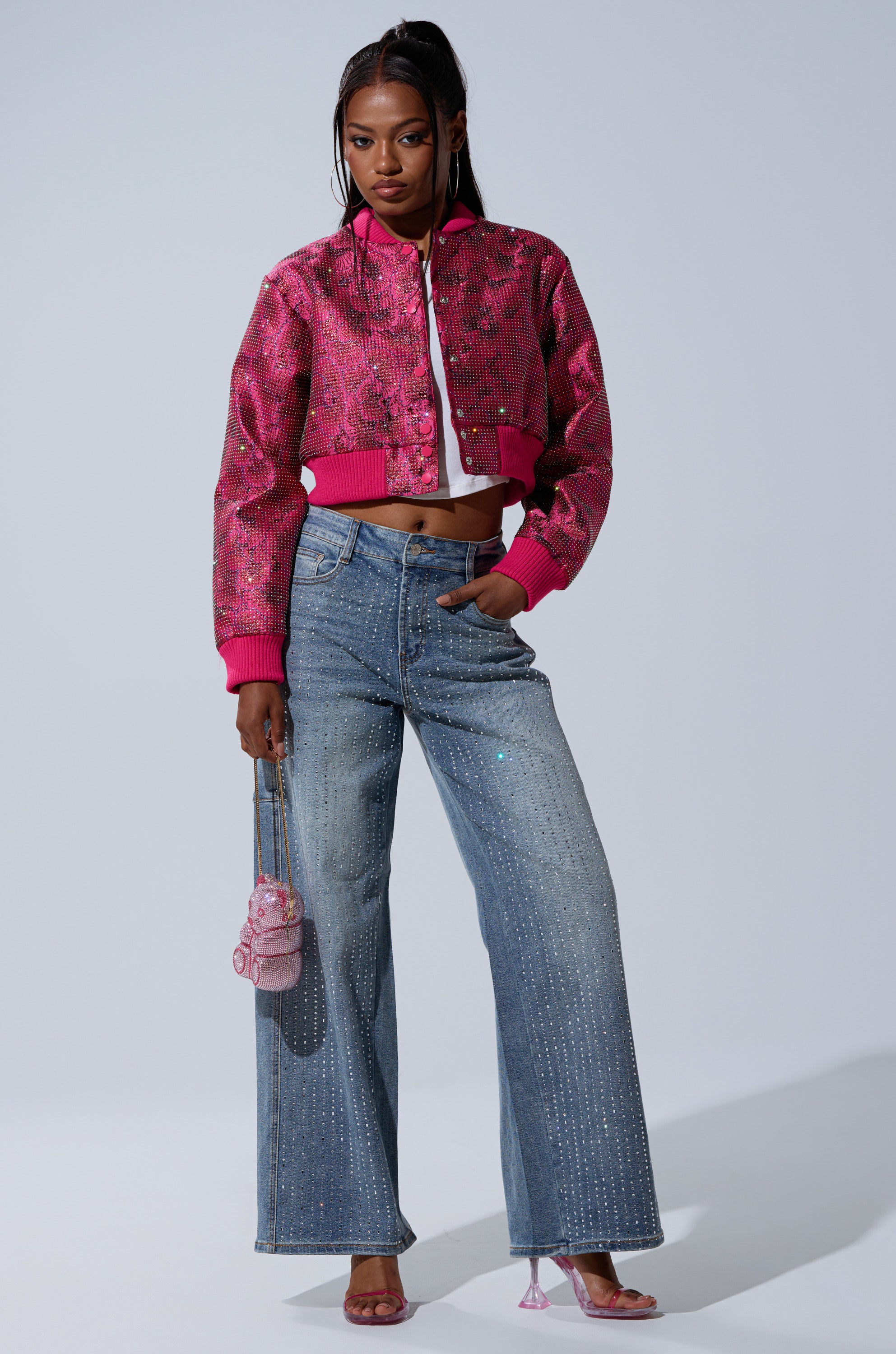 A woman poses against a plain background wearing the FLEUR RHINESTONE BROCADE BOMBER—pink floral with rhinestone details—styled with a white top, wide-leg blue jeans, pink high heels, and a small pink handbag.