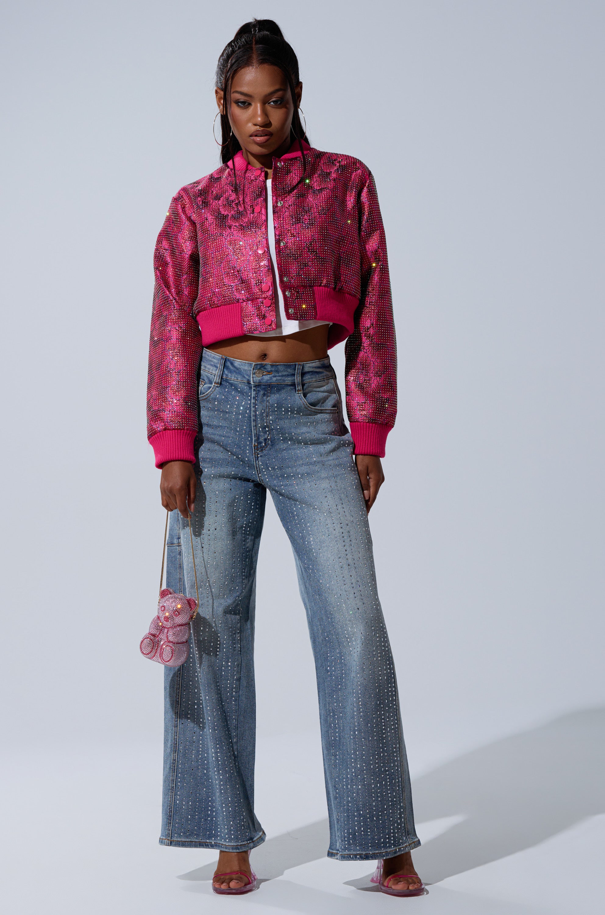A woman wears the FLEUR RHINESTONE BROCADE BOMBER, high-waisted wide-leg blue jeans with rhinestone details, clear heels, and holds a small pink bear-shaped purse, exuding confidence.