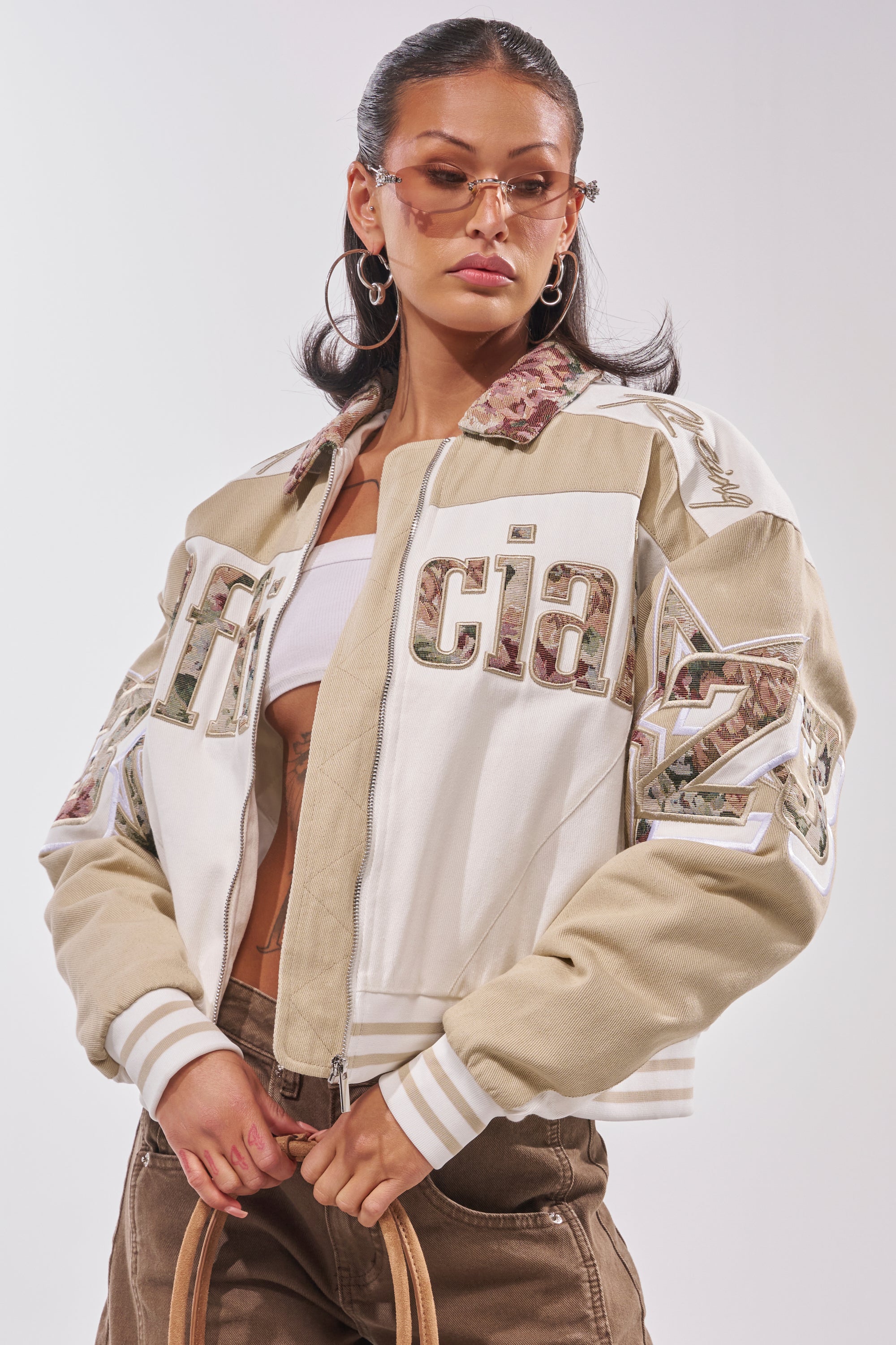 A woman poses confidently in the STILL THE ONE EMBROIDERED BOMBER, styled with brown pants, a white crop top, hoop earrings, tinted sunglasses, and a tan handbag. 1