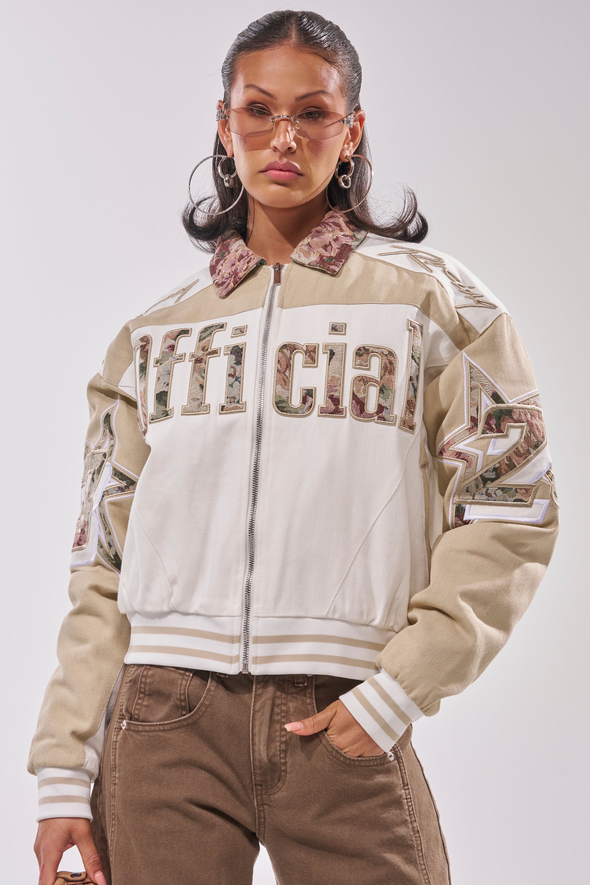 A woman wears the STILL THE ONE EMBROIDERED BOMBER—white and beige with "Official" and "22" details—paired with tinted sunglasses and brown pants, standing against a light background, one hand in her pocket. 2