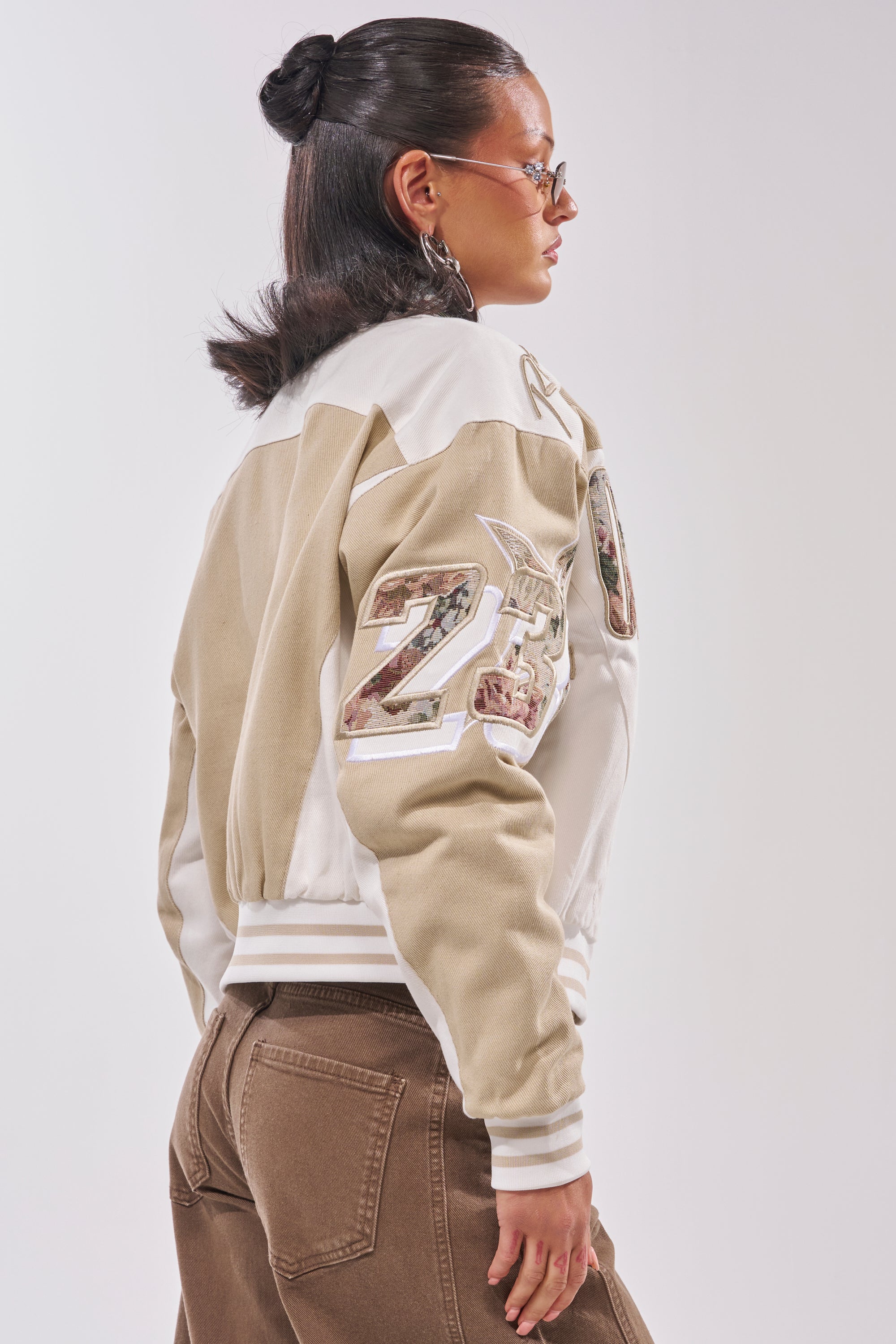A woman with slicked-back hair and sunglasses poses in the STILL THE ONE EMBROIDERED BOMBER, an oversized varsity jacket with large "23" sleeve patches, styled with brown high-waisted pants against a white background. 3