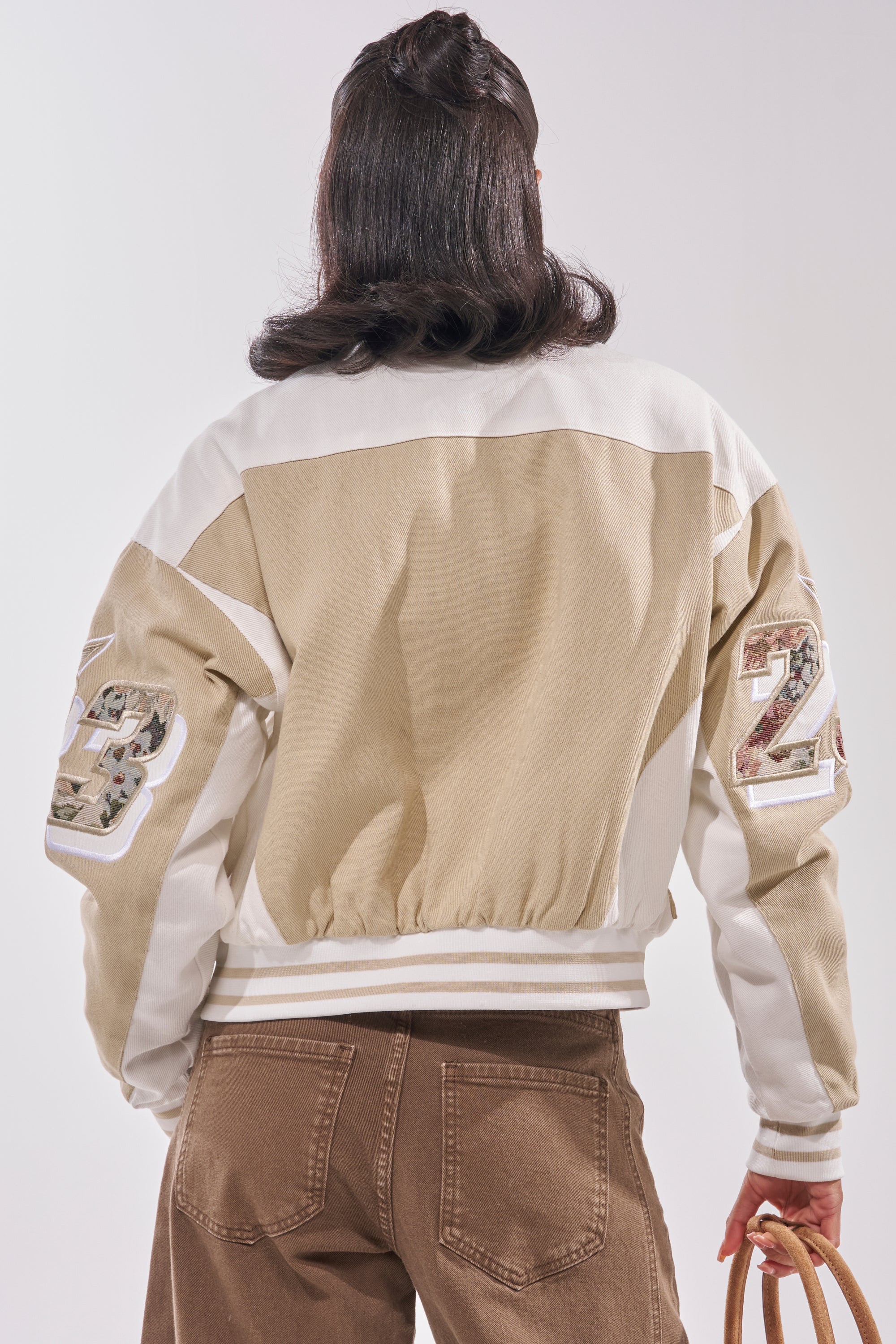 A person stands facing away, wearing the STILL THE ONE EMBROIDERED BOMBER in tan and white with number patches on the sleeves, brown jeans, and holding a light bag. The background is neutral and uncluttered. 4