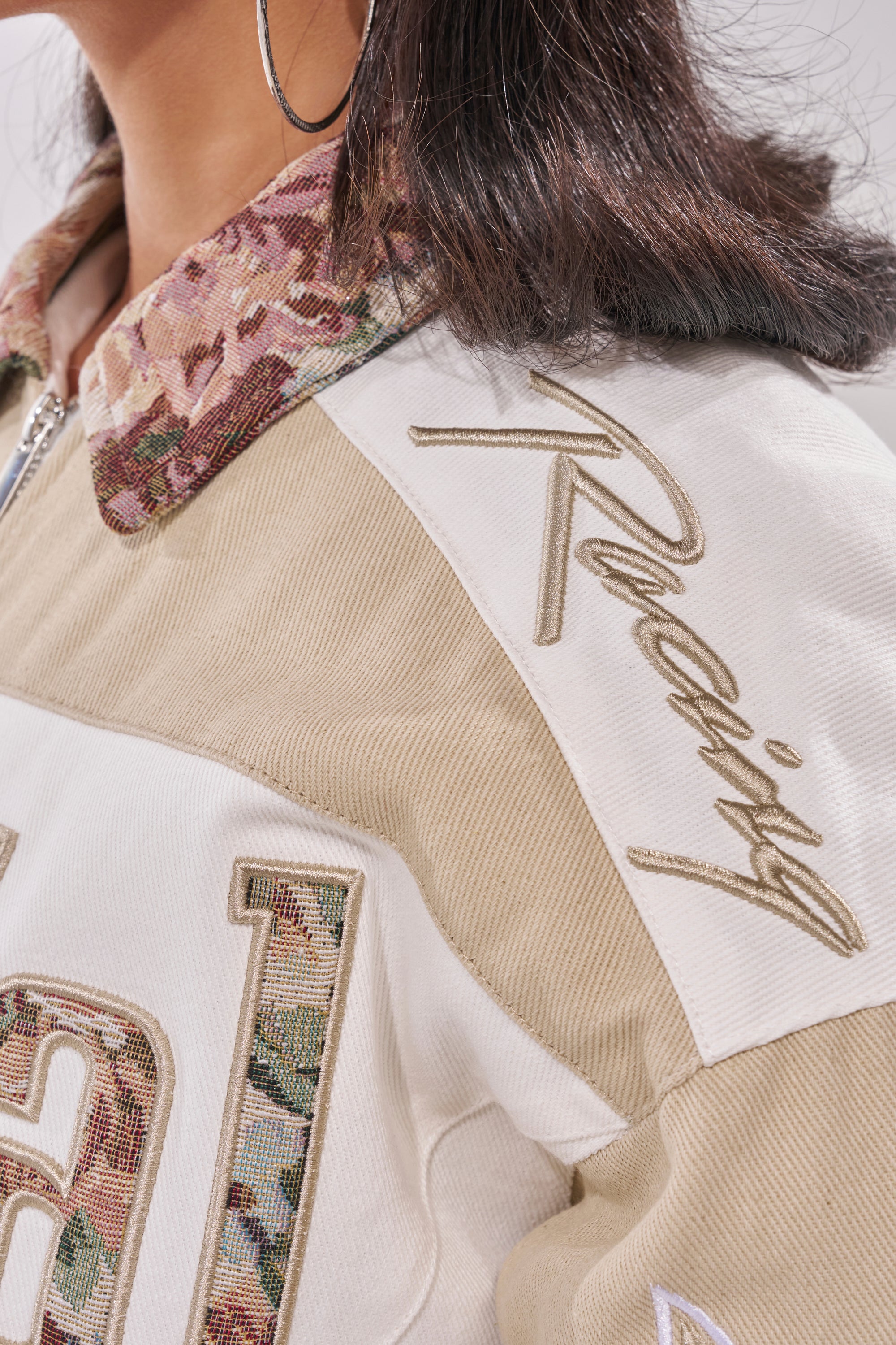 Close-up of a person wearing the STILL THE ONE EMBROIDERED BOMBER, featuring a beige and white colorblock design, floral-patterned collar, and "Racing" text on the shoulder. Paired with straight dark hair and large hoop earring. 8