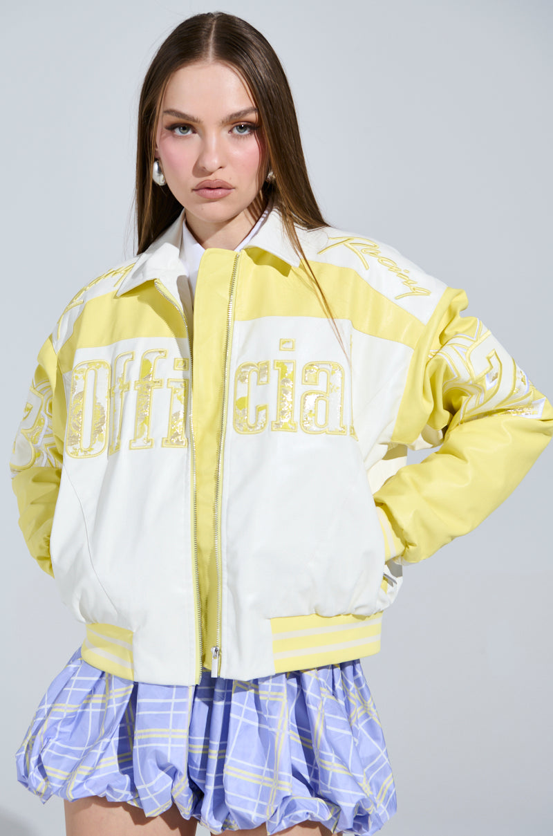 A woman with long brown hair confidently wears a yellow and white UNOFFICIAL RACER BOMBER jacket featuring "Official" on the front. The color block design pairs perfectly with her blue and white plaid skirt against a gray background, highlighting her stylish ensemble.