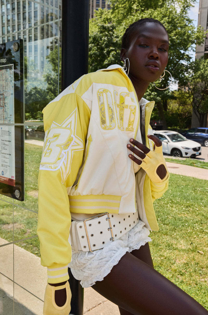 A stylish person stands at a bus stop wearing the UNOFFICIAL RACER BOMBER, lace shorts, fingerless gloves, large hoop earrings, and a studded white belt, with buildings, trees, and cars in the background.