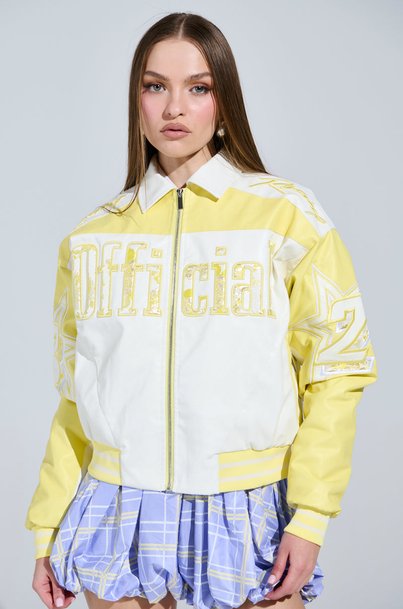 A woman with long brown hair wears a yellow and white UNOFFICIAL RACER BOMBER jacket with "Official" printed on it, paired with a blue and white plaid skirt against a plain gray background.