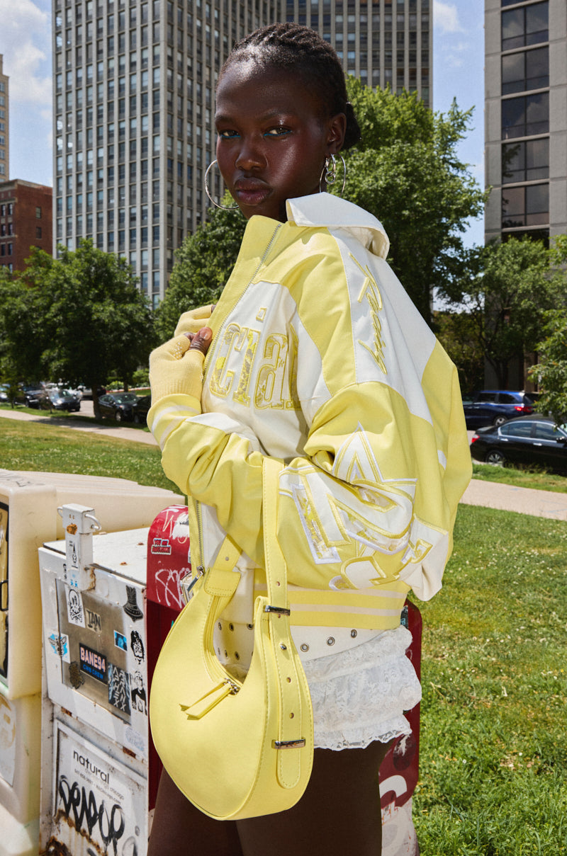 A woman stands outdoors in the city on a sunny day, wearing the UNOFFICIAL RACER BOMBER in yellow and white with white lace shorts, holding a matching yellow bag. Tall buildings and trees appear in the background.