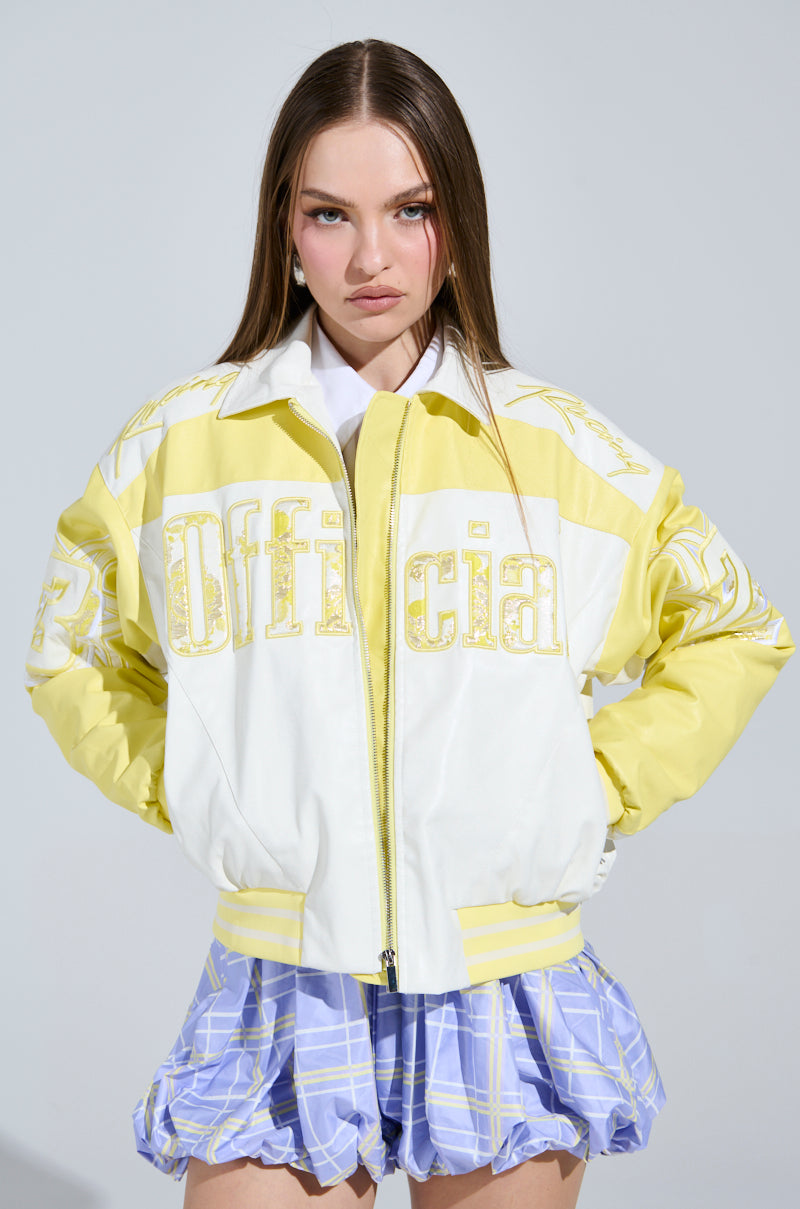 A young woman with long hair stands wearing an "Unofficial Racer Bomber," featuring a yellow and white color block design that complements her light blue plaid skirt. She poses seriously against a plain gray background.