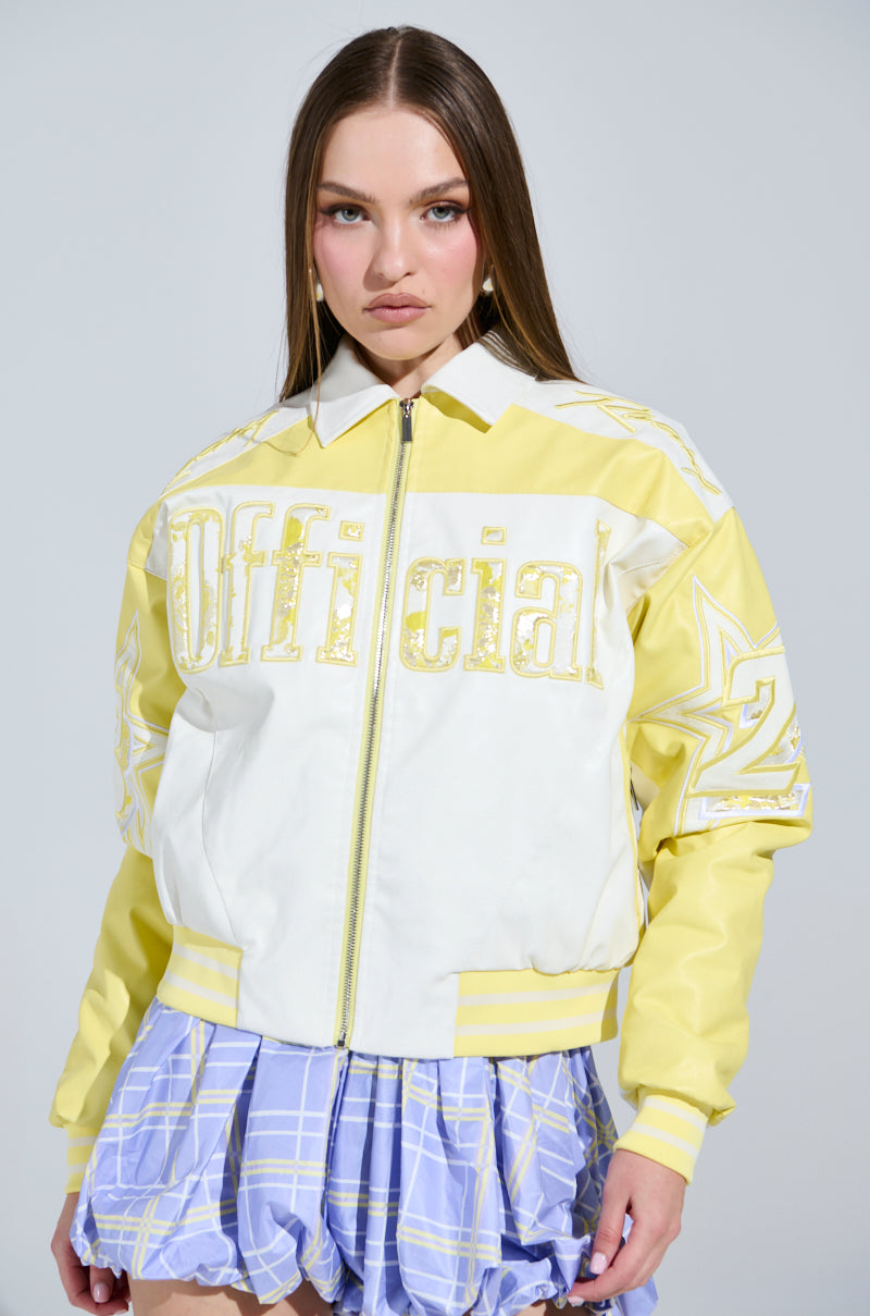 A woman models the UNOFFICIAL RACER BOMBER, a yellow and white jacket with "Official" bolded and stars on the sleeves. The color blocking pairs well with her blue and white plaid skirt. Her long brown hair cascades as she poses against a plain backdrop.