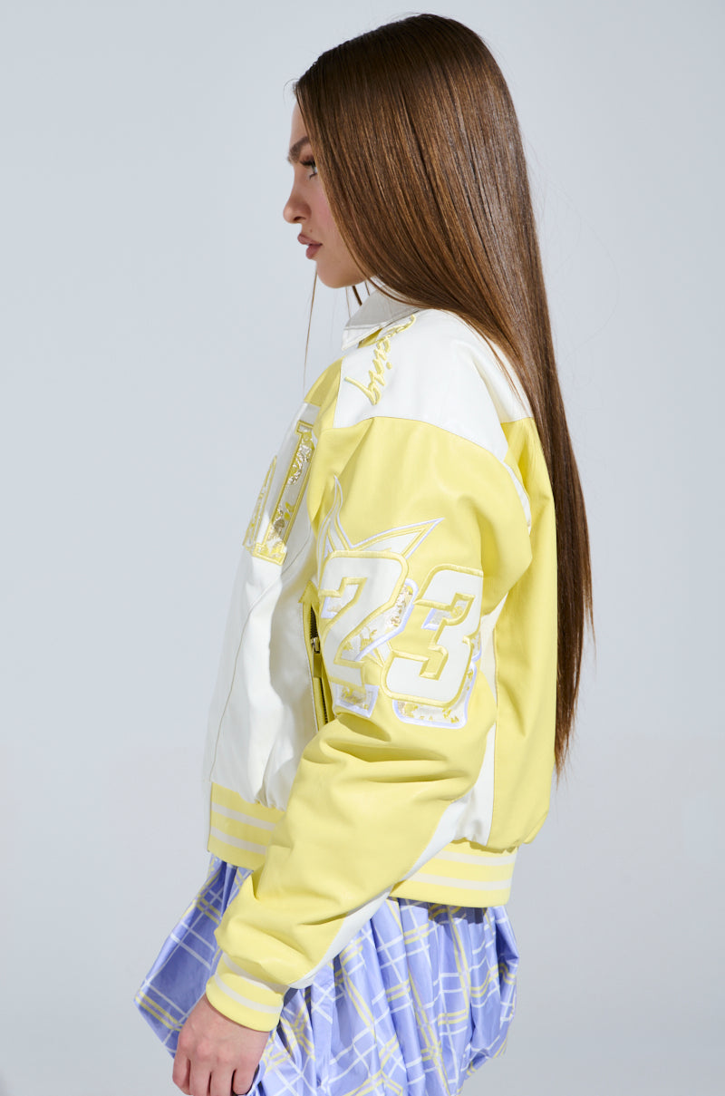 A person with long brown hair wears the UNOFFICIAL RACER BOMBER, showcasing a chic color block design with a large "23" on the back. They're also in a purple plaid skirt, standing sideways against a plain background.