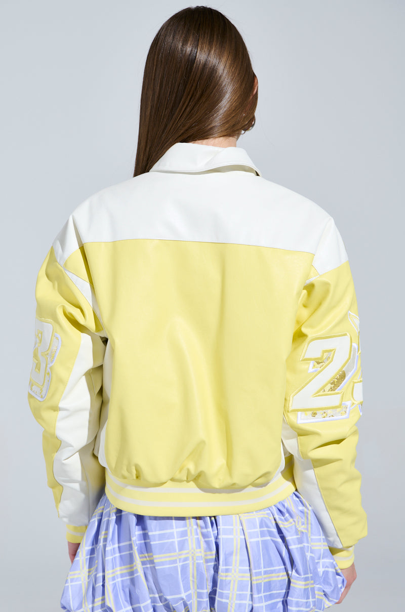 A person with long brown hair wears the UNOFFICIAL RACER BOMBER, a yellow and white jacket featuring "23" on the sleeves in a color block design. They pair it stylishly with a blue and white plaid skirt, standing with their back to the camera.