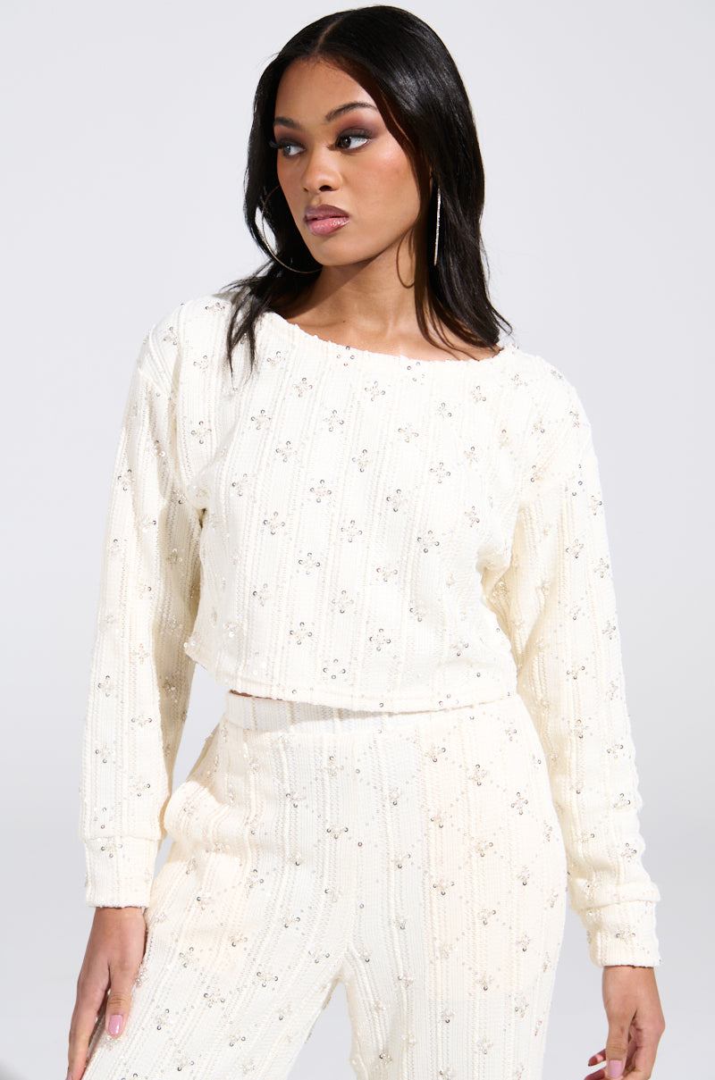 A woman with long dark hair and hoop earrings poses in the SHINE BRIGHT PEARL BOAT NECK SWEATER, a cream-colored patterned knit with sequins, and matching pants. She's standing against a plain background, looking to the side with a neutral expression.