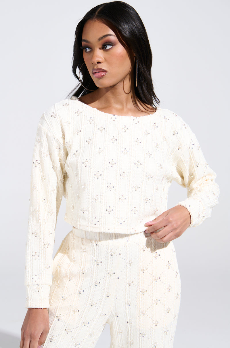 A person with long dark hair wears the SHINE BRIGHT PEARL BOAT NECK SWEATER, a cream-colored, textured top adorned with sequin embellishments and a floral pattern. They stand against a plain white background, looking to the side with one hand on their waist.