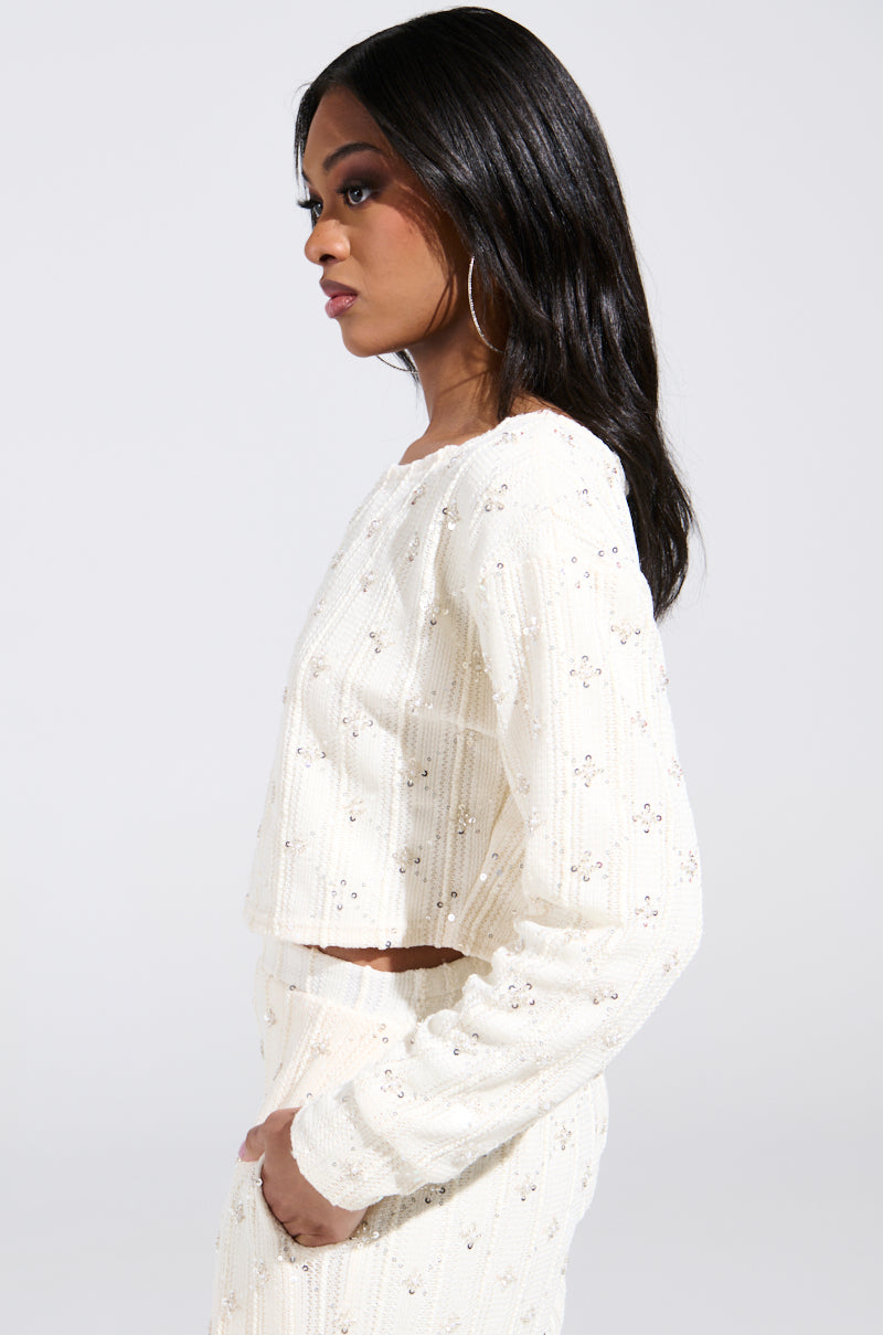 A woman stands against a plain background, looking to the side in hoop earrings and a SHINE BRIGHT PEARL BOAT NECK SWEATER paired with matching pants. Her long, dark hair contrasts with the white textured top adorned with sequin embellishments.