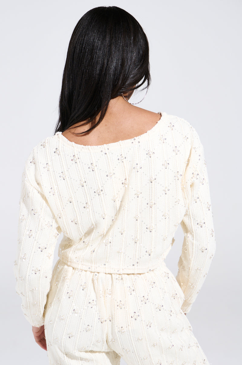 Against a light gray background, a person with long dark hair is showcasing the SHINE BRIGHT PEARL BOAT NECK SWEATER. Adorned with small floral patterns, the cream-colored sweater features a relaxed fit and exudes understated elegance.
