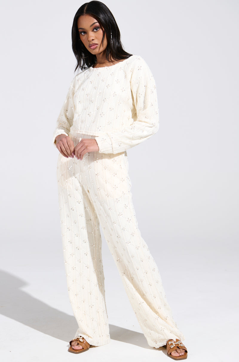 Wearing the SHINE BRIGHT PEARL BOAT NECK SWEATER, a person poses against a gray background. Their outfit features cream-colored, sequin-embellished fabric and long sleeves. With long, dark hair and flat sandals, they look directly at the camera.