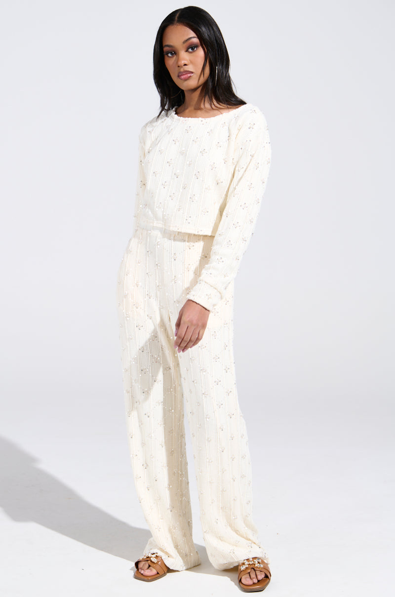 A person stands against a plain backdrop in a cream-colored knit ensemble with subtle embroidery. The look features the elegant SHINE BRIGHT PEARL BOAT NECK SWEATER paired with matching pants and brown sandals.