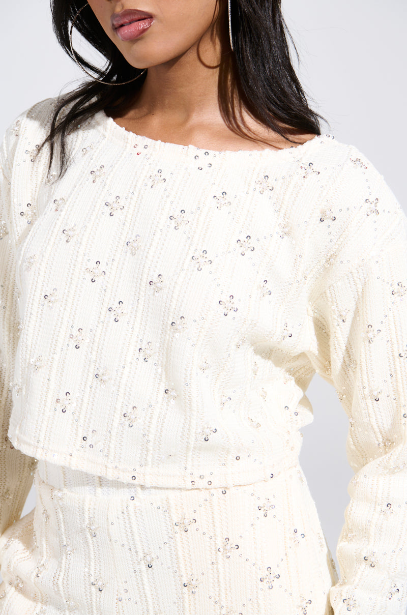 A woman showcases the SHINE BRIGHT PEARL BOAT NECK SWEATER, a white, long-sleeve top with floral patterns and sequin embellishments. The image captures her from shoulders to hips, highlighting her dark hair against a neutral background.