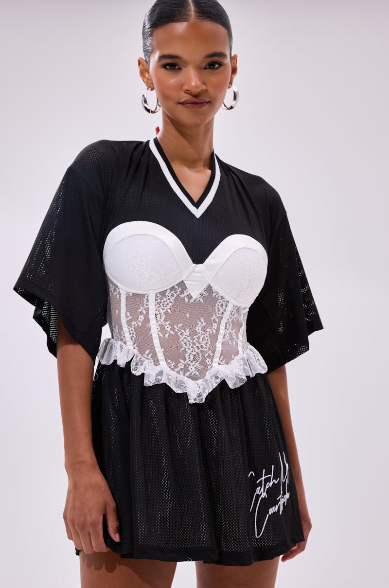 A woman wears the BOUNCE BACK CORSET JERSEY DRESS—black mesh with loose sleeves over a white lace bustier, white V-neckline, and signature graphic near the hem—styled with hoop earrings and slicked-back hair for a chic, sporty look.