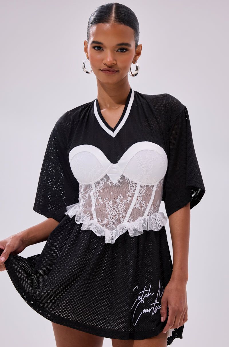 A woman wears the BOUNCE BACK CORSET JERSEY DRESS, a black mesh lace piece with short sleeves layered over a white lace bustier corset and featuring white script text near the hem. She styles her hair back and accessorizes with hoop earrings.
