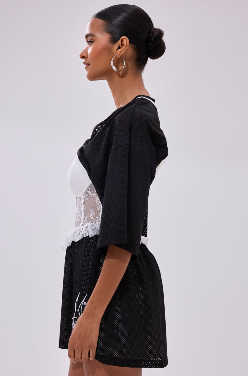 A woman in profile against a plain background wears the BOUNCE BACK CORSET JERSEY DRESS featuring sporty mesh, white lace at the waist, a pleated skirt, and large hoop earrings. Her hair is styled in a neat bun.