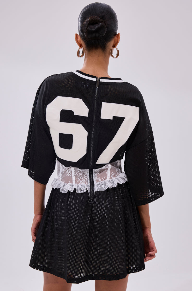 A woman with her hair in a bun and hoop earrings stands with her back to the camera, wearing the BOUNCE BACK CORSET JERSEY DRESS featuring sporty mesh fabric, white lace at the waist, and the number 67 in white on the back.