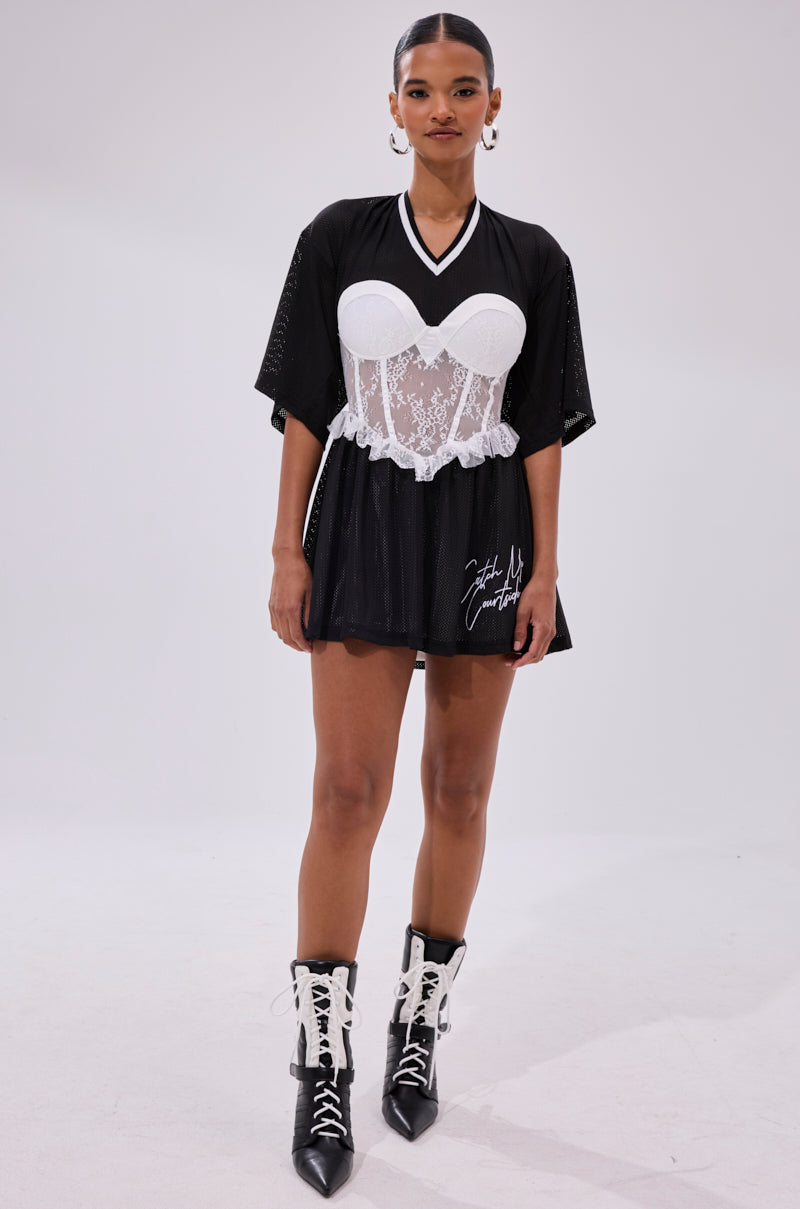A woman stands confidently in the BOUNCE BACK CORSET JERSEY DRESS, featuring black fabric and white lace-up details, paired with hoop earrings and black boots with white accents against a plain background.