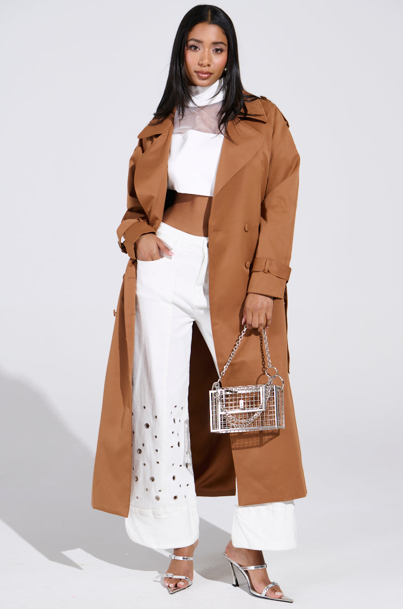 A woman wears the NEW YORK MINUTE TRENCH IN BROWN over a white, sheer-paneled crop top and wide-leg pants with cut-out designs. She holds a transparent handbag and dons silver heels, confidently posing against a plain background. 0