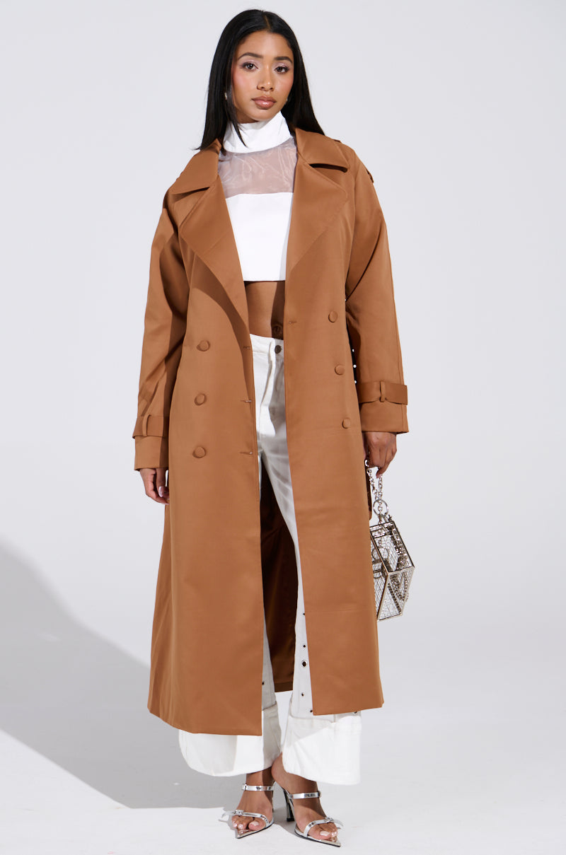 A person models an oversized, brown New York Minute Trench with an adjustable waistbelt over a white crop top and pants, holding a patterned handbag. They wear silver heels against a plain, light-colored background. 1