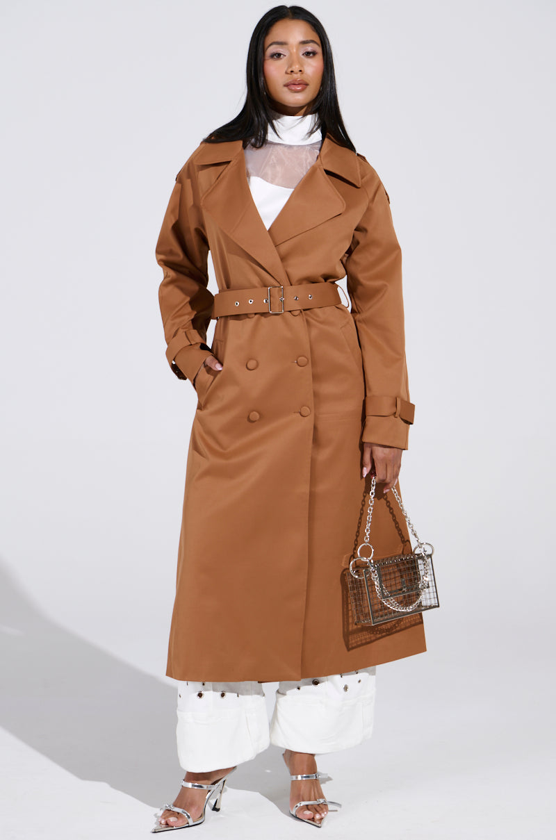 A person confidently poses against a plain gray background, wearing the NEW YORK MINUTE TRENCH IN BROWN with an adjustable waistbelt over a white outfit. They hold a clear handbag and wear silver open-toe heels. 2