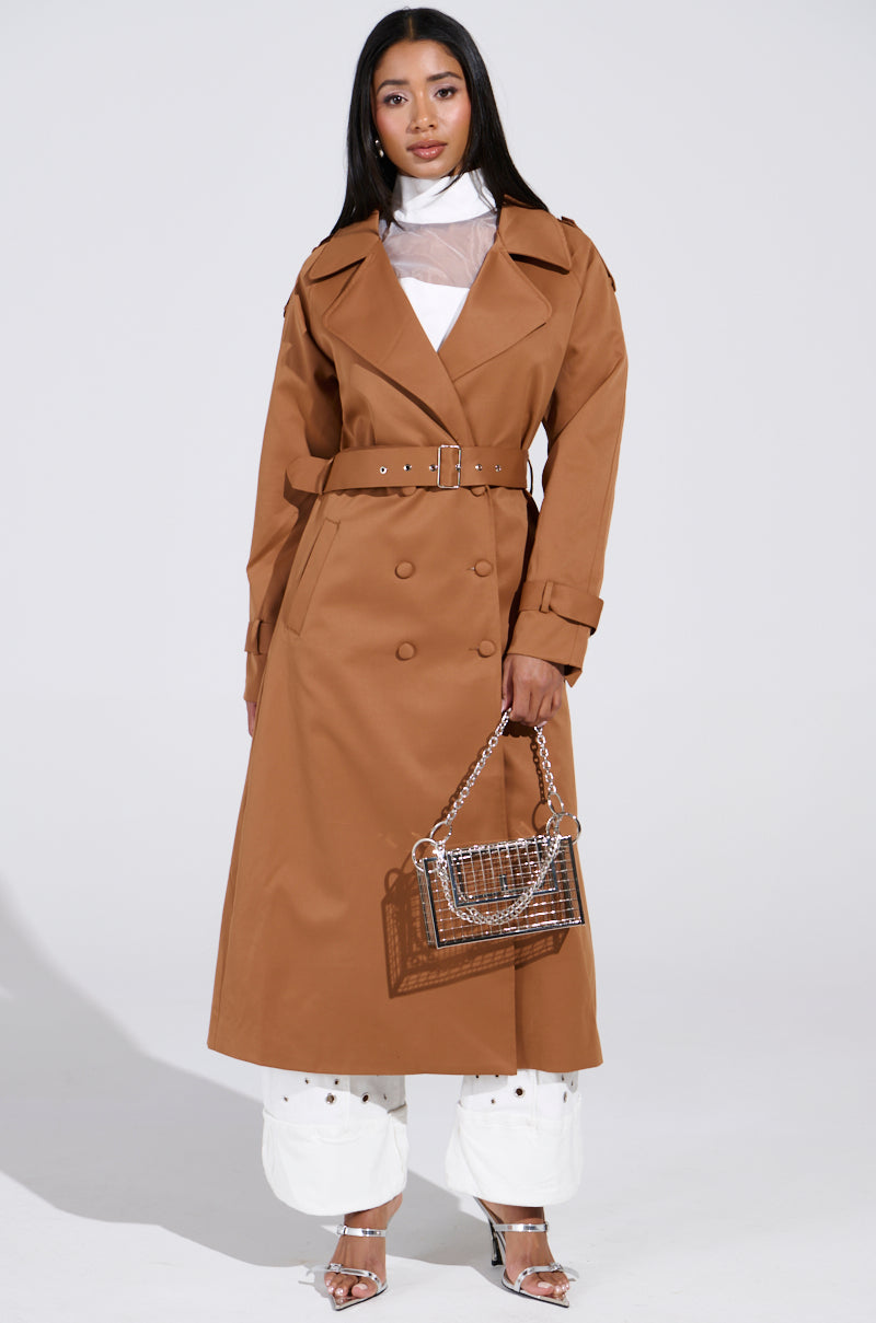 A woman confidently stands in the NEW YORK MINUTE TRENCH IN BROWN with an adjustable waistbelt. She carries a clear handbag, wears silver high heels that complement her long, straight hair beautifully against the plain gray backdrop. 3