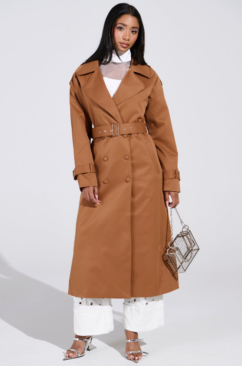 A woman exudes confidence in a New York Minute Trench in brown, paired with a sheer white turtleneck, white pants, and silver heels. Her transparent handbag complements the adjustable waistbelt of the trench against the plain, well-lit backdrop. 4