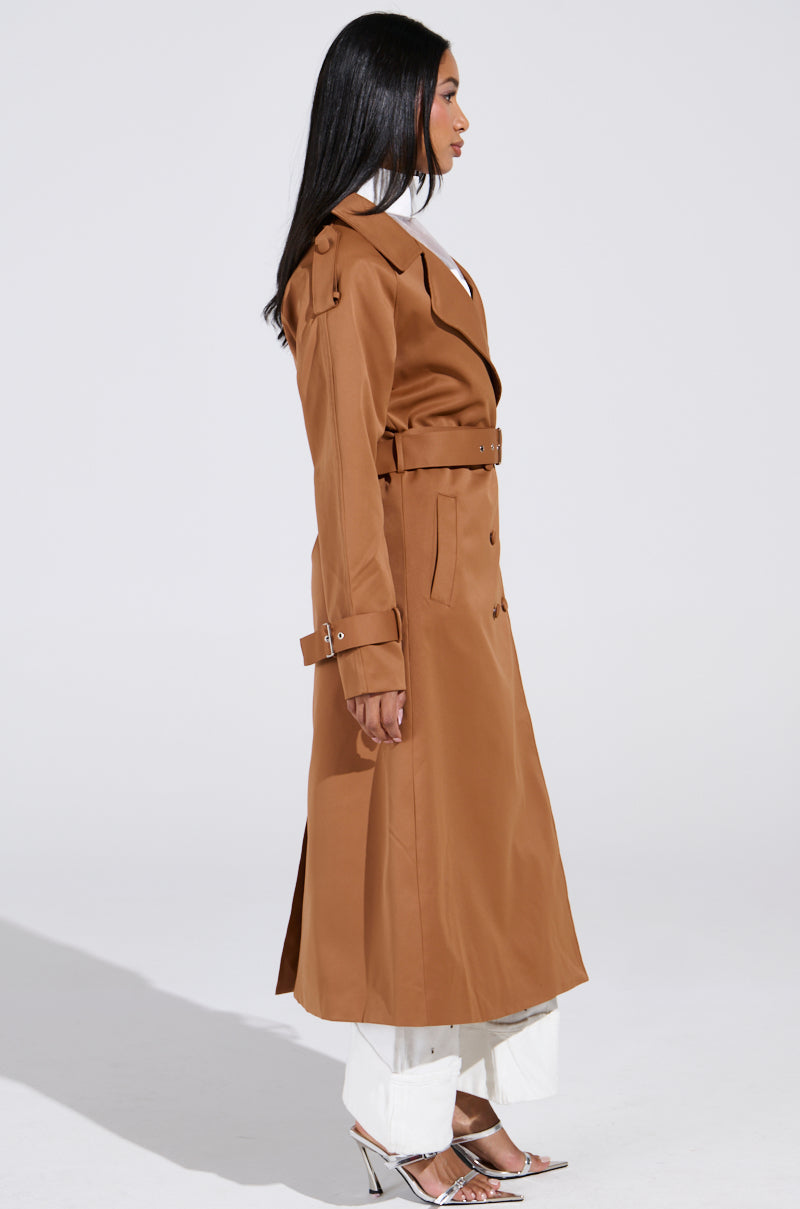 A person with long black hair stands in profile, highlighting the oversized New York Minute Trench in brown, complete with an adjustable waistbelt and button details. They pair it with white pants and silver heels against a plain white backdrop. 5