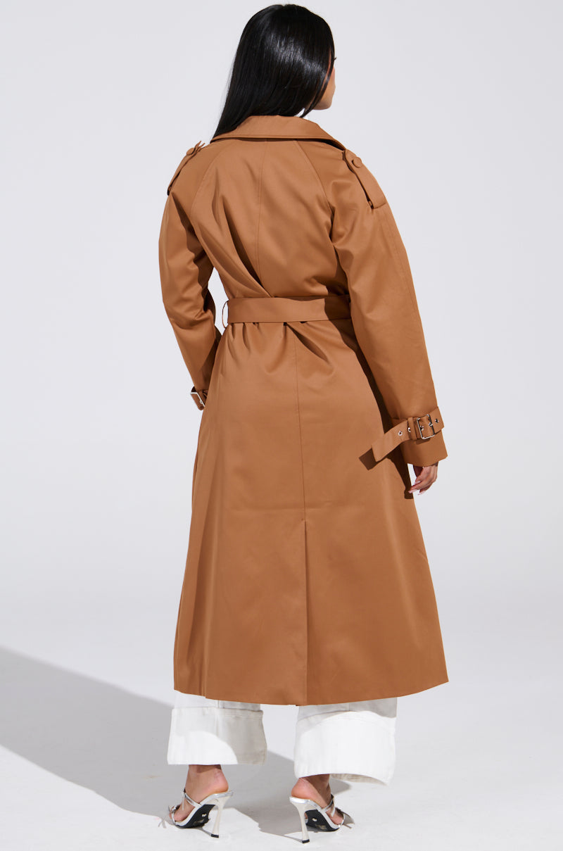 A person with long black hair wears the NEW YORK MINUTE TRENCH IN BROWN, featuring an adjustable waistbelt. They pair it with white pants and open-toe high heels, standing against a plain white background, facing away from the camera. 6