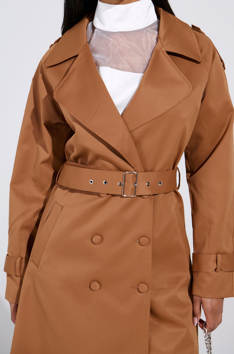 A person wears the oversized NEW YORK MINUTE TRENCH IN BROWN, featuring button details and an adjustable waistbelt, over a white high-collared shirt. 7