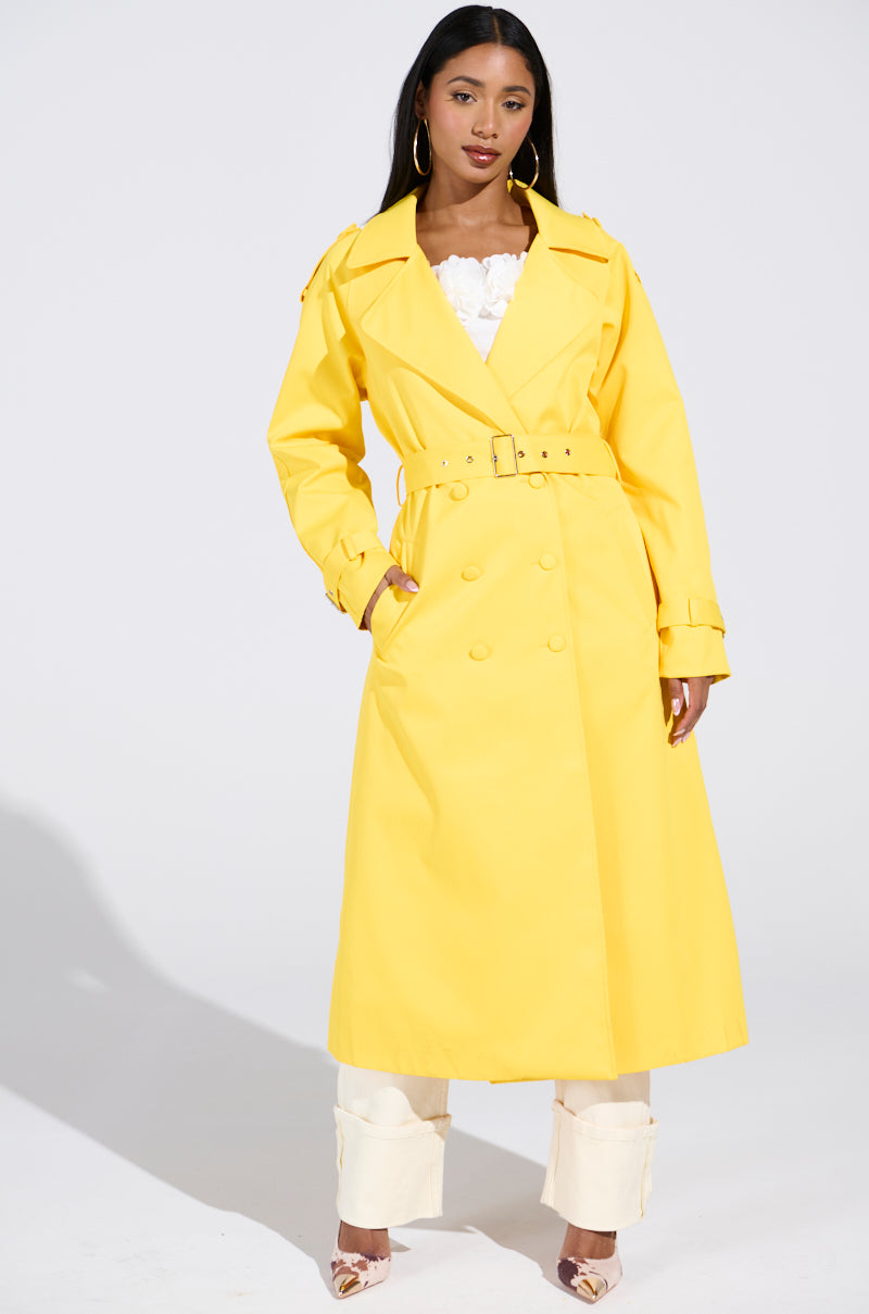 Wearing the vibrant NEW YORK MINUTE TRENCH IN YELLOW with an oversized fit and satin lining, a person stands confidently. They pair it with light-colored pants and heels against a plain backdrop, highlighting the striking coat.