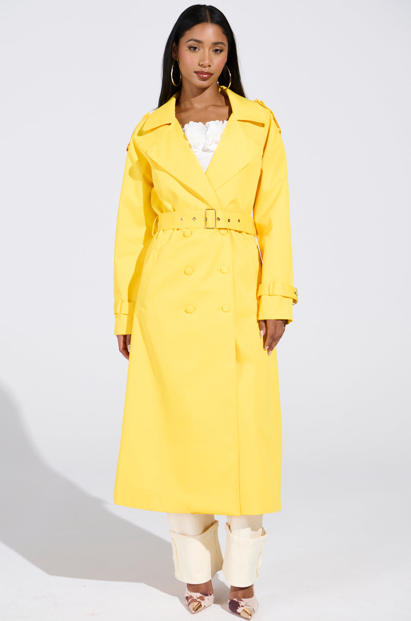 A person with long hair is wearing an oversized NEW YORK MINUTE TRENCH IN YELLOW, satin-lined for luxury, over a white outfit. They stand against a plain background. The coat has a belt and double-breasted buttons.
