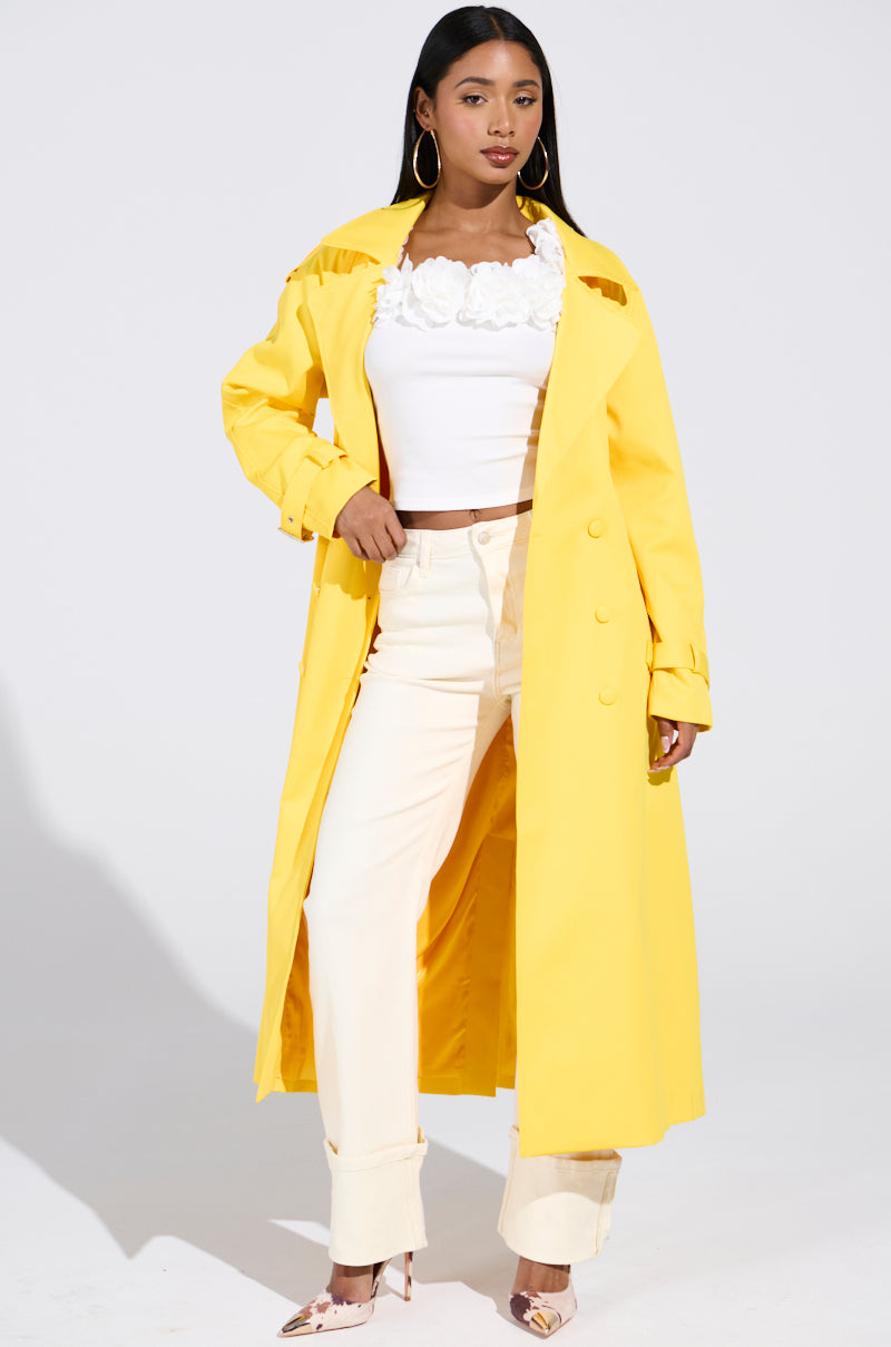 A woman confidently poses in the NEW YORK MINUTE TRENCH IN YELLOW over a white top and light pants. Her long hair cascades over the satin-lined coat, accentuating her large round earrings against the plain white background.