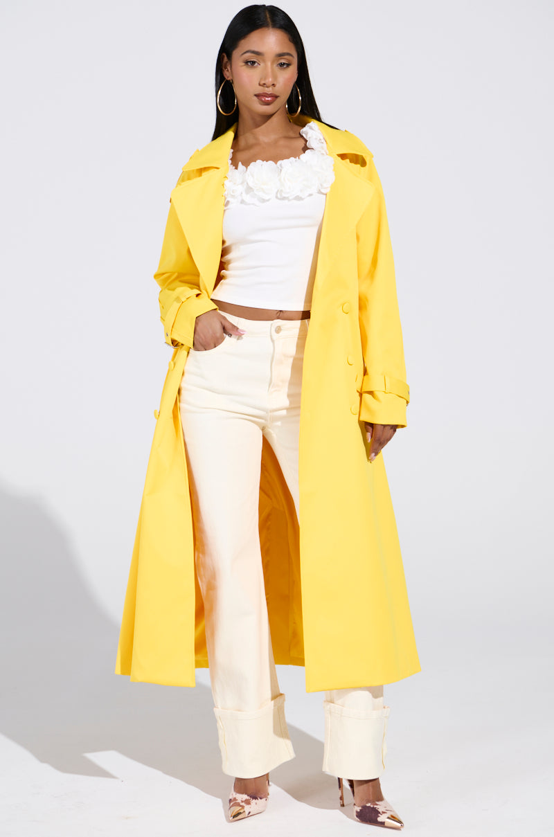 A person poses confidently in an oversized NEW YORK MINUTE TRENCH IN YELLOW over a white flower-embellished top and cream pants with rolled cuffs, wearing hoop earrings and pointed patterned shoes, against a plain background.