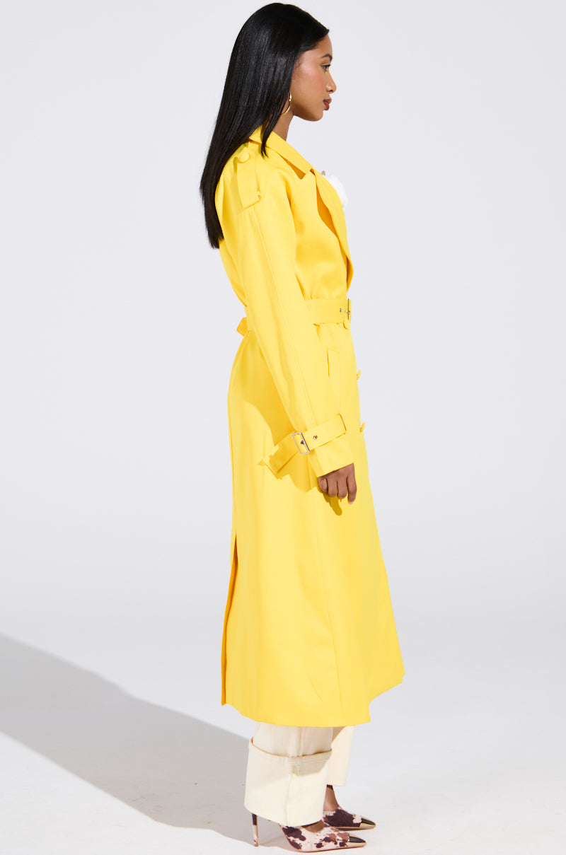 A person with long hair stands in profile wearing the oversized-fit New York Minute Trench in yellow, featuring belt and button details. The satin-lined coat complements their white pants and pointed heels against the plain white background.