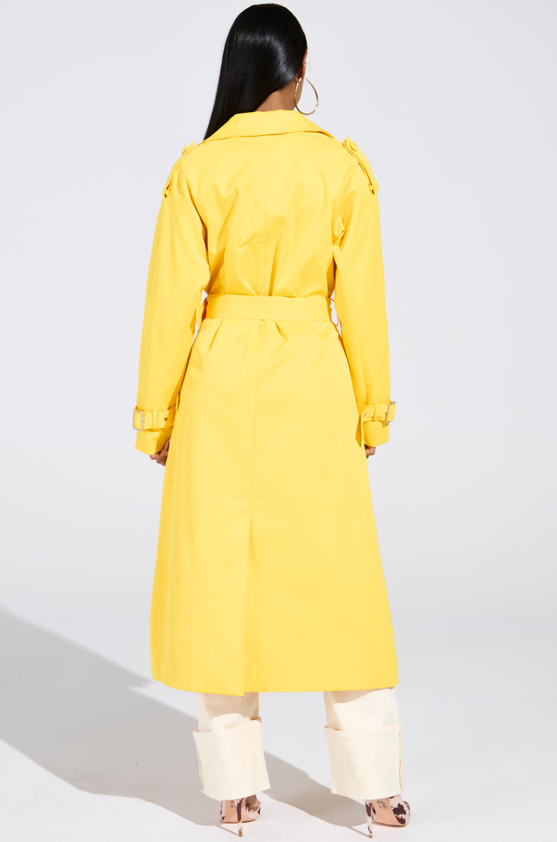 A person with long dark hair wears the NEW YORK MINUTE TRENCH IN YELLOW, an oversized bright trench with a double-breasted closure, paired with light-colored pants. They stand with their back to the camera against a plain background.