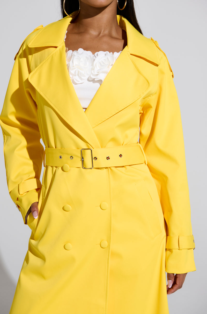 Dressed in the NEW YORK MINUTE TRENCH IN YELLOW, someone stylishly flaunts its oversized fit and large lapels, with satin lining peeking underneath. The trench is cinched at the waist over a floral-detailed white top, as they pose confidently with one hand in their pocket.