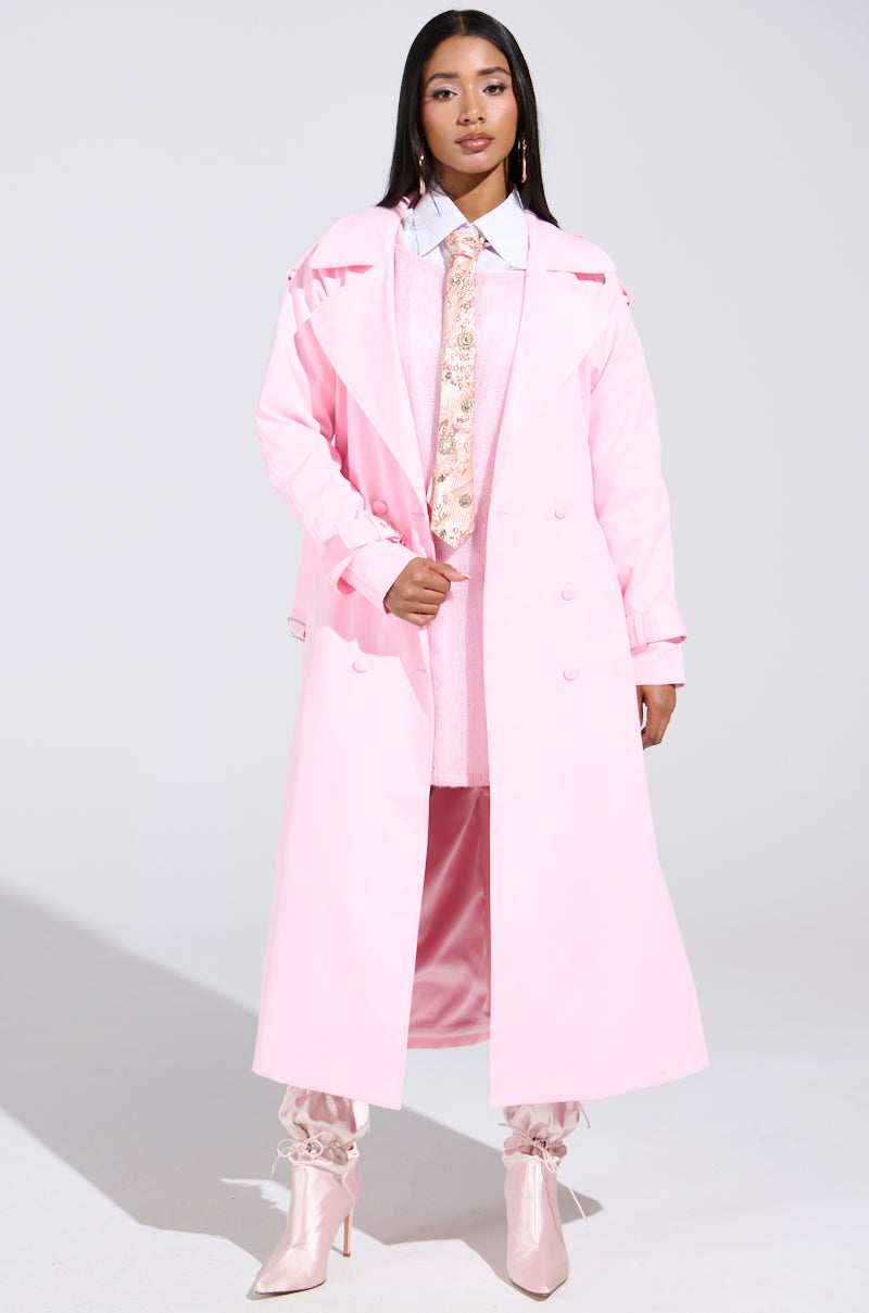 A confident individual wears the PRETTY IN PINK TRENCH over a matching outfit, complete with a patterned tie and light pink ruffled high-heeled boots. A plain, well-lit background highlights the vibrant charm of their feminine spring jacket ensemble.