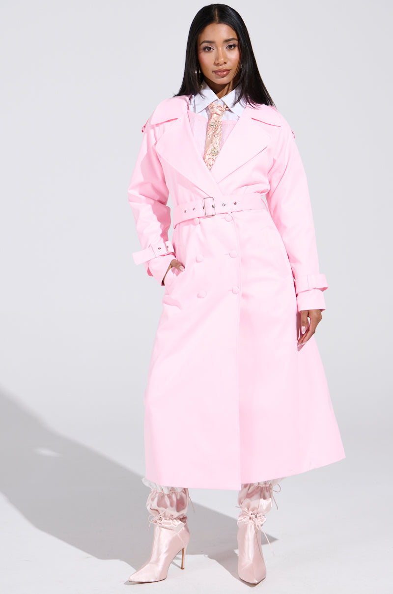 A woman exudes confidence in the PRETTY IN PINK TRENCH with matching pants, a white collared shirt, sparkly tie, and high-heeled boots. The plain white background enhances her pastel look, crafting an ideal feminine spring style.