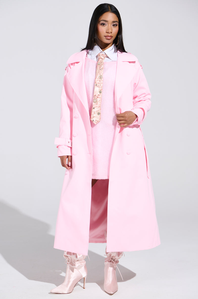 A person stands confidently in the PRETTY IN PINK TRENCH over a light sweater and floral tie, paired with shiny pointed pink boots and white socks against a plain light gray background.
